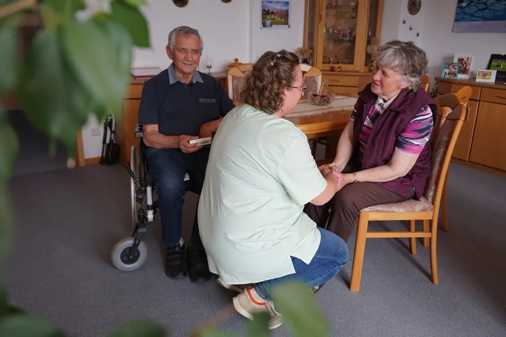 Ein älterer Mann im Rollstuhl und eine ältere Frau sitzen in einem gemütlichen Zimmer. Eine Pflegerin der Ambulanten Dienste in grüner Uniform ist mit dabei. Das Zimmer ist mit Holzmöbeln und dekorativen Elementen an der Wand ausgestattet.
