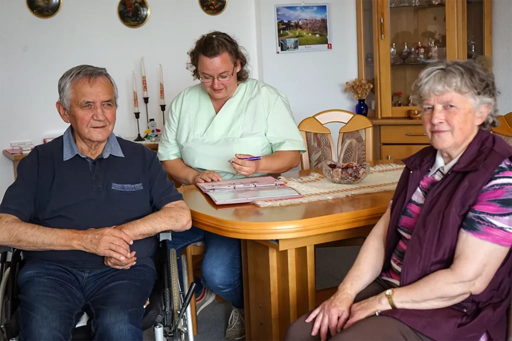 Ein älterer Mann im Rollstuhl und eine ältere Frau sitzen in einem gemütlichen Zimmer. Eine Pflegerin der Ambulanten Dienste in grüner Uniform sitzt an einem Tisch hinter ihnen. Das Zimmer ist mit Holzmöbeln und dekorativen Tellern an der Wand ausgestattet.