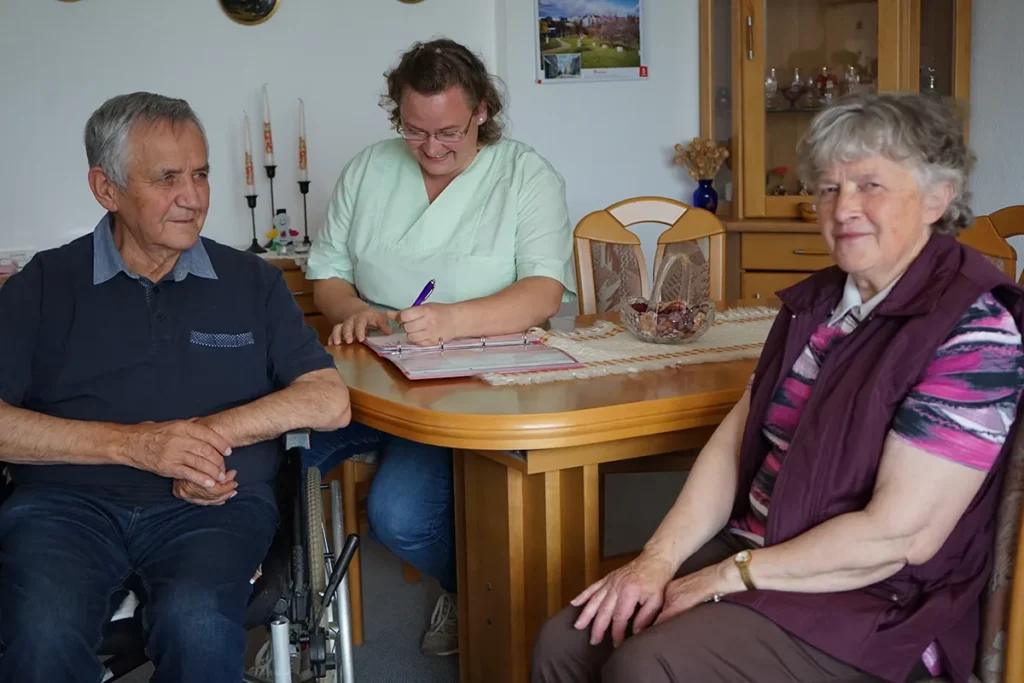 Ein älterer Mann im Rollstuhl und eine ältere Frau sitzen in einem gemütlichen Zimmer. Eine Pflegerin der Ambulanten Dienste in grüner Uniform ist mit dabei. Das Zimmer ist mit Holzmöbeln und dekorativen Elementen an der Wand ausgestattet.