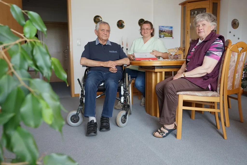 Ein älterer Mann im Rollstuhl und eine ältere Frau sitzen in einem gemütlichen Zimmer. Eine Pflegerin der Ambulanten Dienste in grüner Uniform ist mit dabei. Das Zimmer ist mit Holzmöbeln und dekorativen Elementen an der Wand ausgestattet.