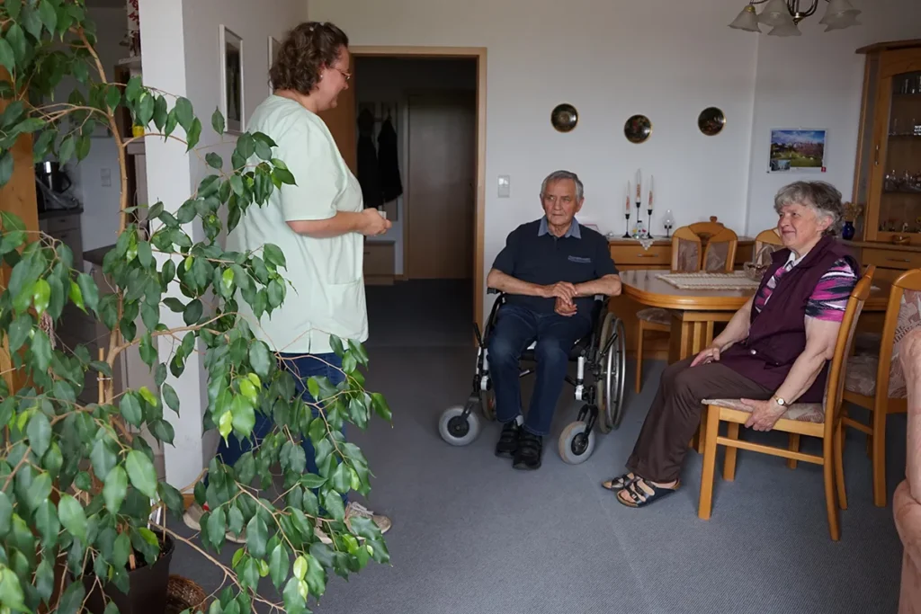 Ein älterer Mann im Rollstuhl und eine ältere Frau sitzen in einem gemütlichen Zimmer. Eine Pflegerin der Ambulanten Dienste in grüner Uniform ist mit dabei. Das Zimmer ist mit Holzmöbeln und dekorativen Elementen an der Wand ausgestattet.