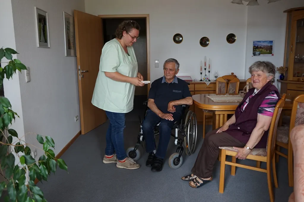Ein älterer Mann im Rollstuhl und eine ältere Frau sitzen in einem gemütlichen Zimmer. Eine Pflegerin der Ambulanten Dienste in grüner Uniform ist mit dabei. Das Zimmer ist mit Holzmöbeln und dekorativen Elementen an der Wand ausgestattet.