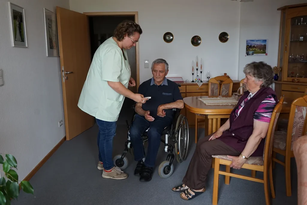 Ein älterer Mann im Rollstuhl und eine ältere Frau sitzen in einem gemütlichen Zimmer. Eine Pflegerin der Ambulanten Dienste in grüner Uniform ist mit dabei. Das Zimmer ist mit Holzmöbeln und dekorativen Elementen an der Wand ausgestattet.