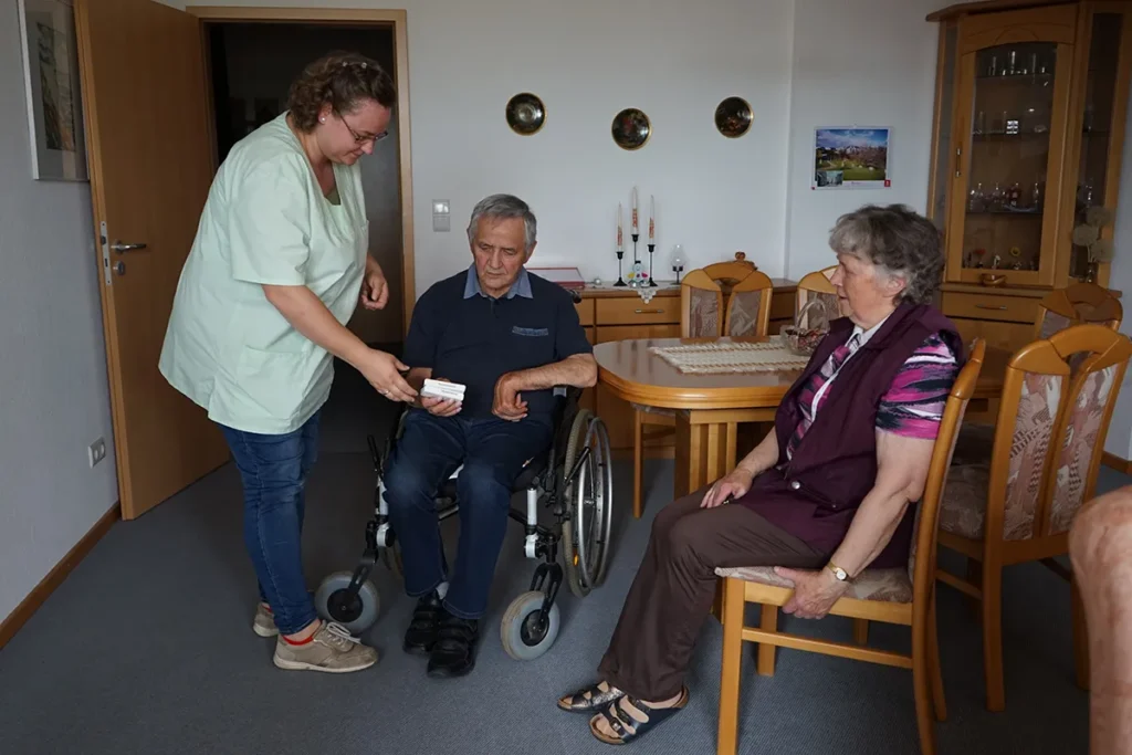 Ein älterer Mann im Rollstuhl und eine ältere Frau sitzen in einem gemütlichen Zimmer. Eine Pflegerin der Ambulanten Dienste in grüner Uniform ist mit dabei. Das Zimmer ist mit Holzmöbeln und dekorativen Elementen an der Wand ausgestattet.