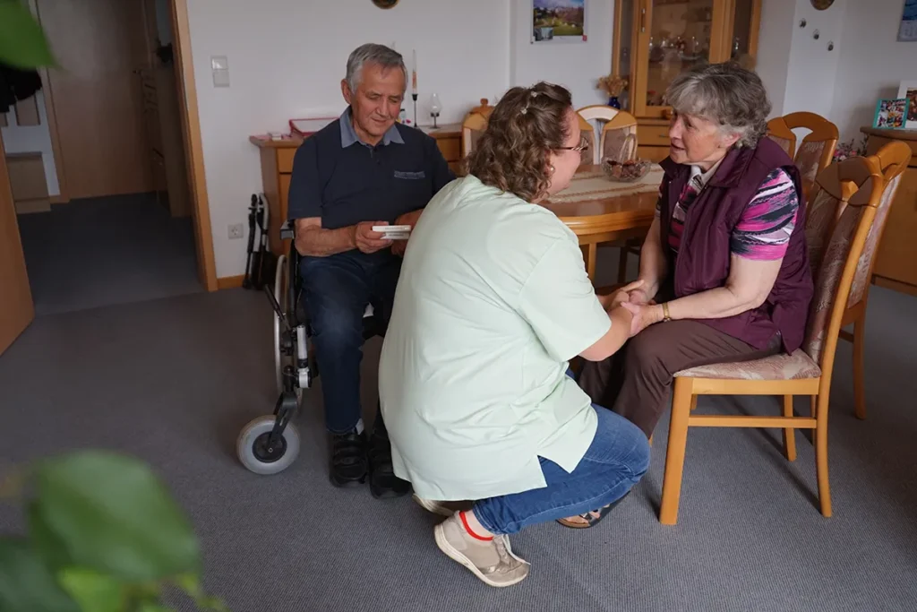 Ein älterer Mann im Rollstuhl und eine ältere Frau sitzen in einem gemütlichen Zimmer. Eine Pflegerin der Ambulanten Dienste in grüner Uniform ist mit dabei. Das Zimmer ist mit Holzmöbeln und dekorativen Elementen an der Wand ausgestattet.
