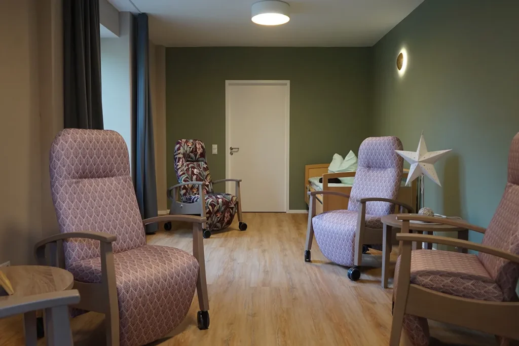 In einem gemütlichen Zimmer der Tagespflege stehen vier gepolsterte Sessel, ein Bett mit Bettwäsche, eine große weiße Sternendekoration, ein Holzboden und eine beruhigende grüne Akzentwand.